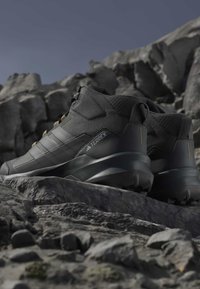 Μαύρα ορειβατικά παπούτσια Adidas Terrex τοποθετημένα σε βραχώδες έδαφος με ορεινό φόντο κάτω από καθαρό ουρανό.