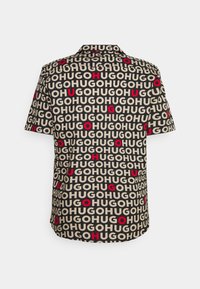Chemise noire à manches courtes avec motif répété de texte beige "HUGO" et lettres rouges placées aléatoirement au dos.