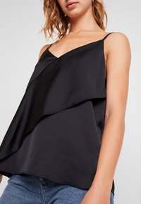 Svart satintopp med V-ringning, med lagerdesign och tunna remmar, i kombination med blå denimunderdelar. Slät textur och flödande form.