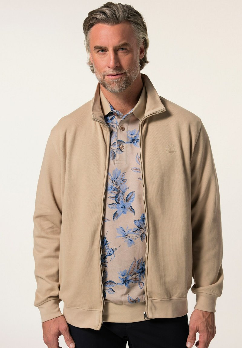 Giacca beige con zip e texture a coste, abbinata a un polo leggera con motivo floreale nei toni del blu e del beige. Il modello ha capelli grigi di lunghezza media.