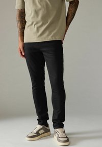 Zwarte skinny jeans met een gladde textuur, gecombineerd met een licht olijfgroen t-shirt en grijze sneakers met beige accenten en veters.