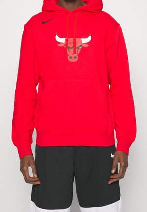 Osoba nosząca czerwoną bluzę z kapturem z logo Chicago Bulls oraz czarne spodenki Nike z białym logo Nike na lewej nodze.