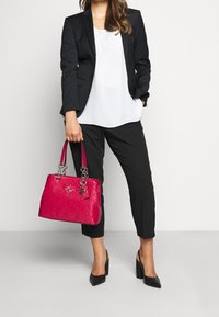 Sac à main rose vif en cuir embossé, avec garnitures argentées, deux poignées et un porte-clés. Porté avec un costume noir et des talons.