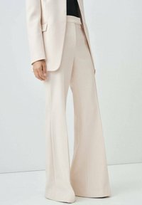 Persona che indossa pantaloni a gamba larga di colore crema e un blazer abbinato con una maglietta nera, in piedi contro uno sfondo chiaro e semplice.