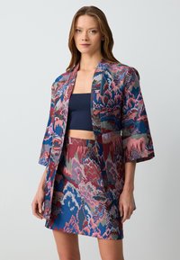 Mehrfarbige kimonoartige Jacke aus strukturiertem Stoff mit einem abstrakten Muster in Rot, Blau und Grün, kombiniert mit einem passenden Rock.