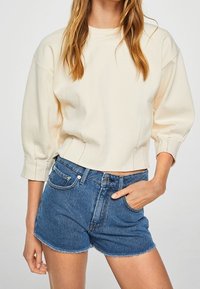 Sudadera recortada color crema con mangas abullonadas, combinada con pantalones cortos de mezclilla azul de cintura alta con el dobladillo deshilachado y cinco bolsillos.