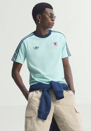 Giovane con maglietta Adidas azzurro chiaro con logo e tre strisce sulle maniche, pantaloni cargo beige e felpa blu marina legata in vita.
