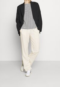 Black blazer, white and black striped shirt, beige trousers, and white sneakers. Casual fit with a structured jacket and soft textures.