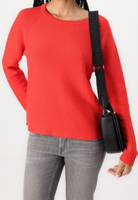 Pull en maille rouge avec un col arrondi et des poignets côtelés, associé à un jean gris et un sac bandoulière noir, avec une sangle à logo.