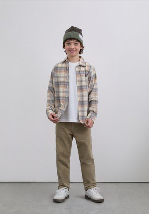 Camisa de cuadros abotonada en beige, azul y naranja, llevada sobre una camiseta blanca, combinada con pantalones caqui y zapatillas blancas. Gorro de punto verde oscuro.