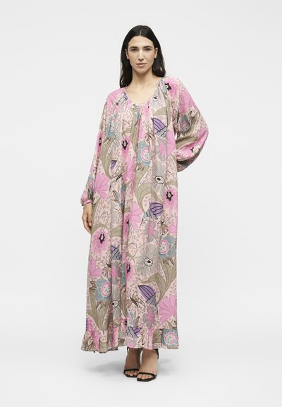 Γυναίκα με μακρύ, φαρδύ φόρεμα με λουλούδια σε ροζ, μοβ και μπεζ αποχρώσεις, όρθια με ένα χέρι στη μέση και μαύρα πέδιλα με τακούνι.