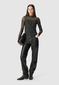 Donna che indossa una blusa trasparente a maniche lunghe con stampa animale, pantaloni di pelle nera, stivali neri alla caviglia, con una pochette nera in mano, in piedi su uno sfondo bianco.