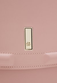 Fermoir rectangulaire en métal doré avec logo gravé sur une surface en cuir rose doux avec détails de couture visibles.
