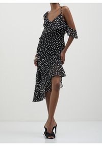 Robe noire à pois avec volants superposés, épaules dénudées, ourlet asymétrique et tissu léger. Portée avec des sandales à talons noires.