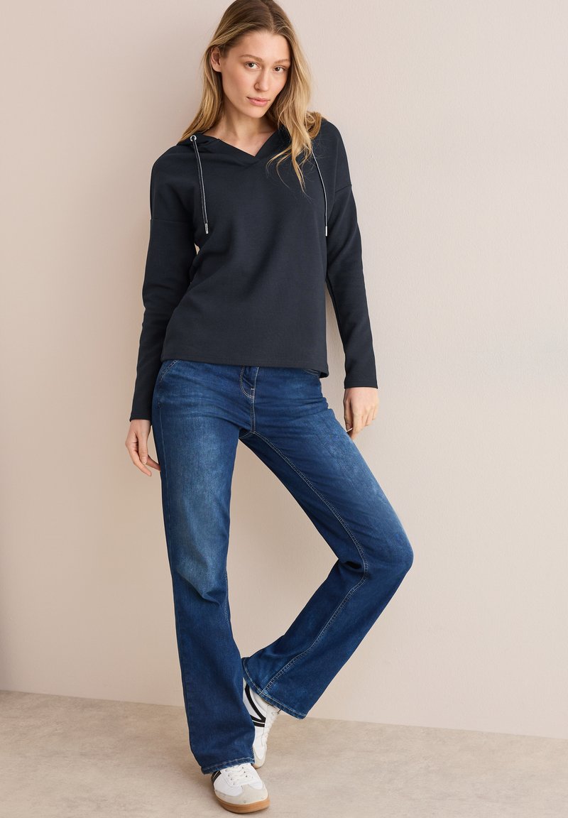 Marineblå hættetrøje med snoretræk, parret med mørkeblå lige jeans og hvide sneakers. Baggrunden er i en blød neutral tone.