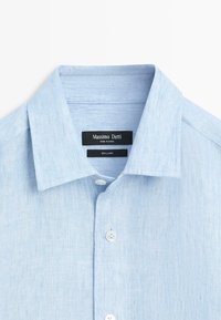Chemise en lin bleu clair avec col ouvert, boutons blancs et une étiquette noire indiquant "Massimo Dutti" et "100 % lin" à l'intérieur du col.