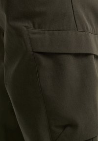 Dunkelolivgrüne Hose aus glattem Stoff, mit einer frontalen Tasche mit Klappe und detaillierter Naht entlang der Nähte.