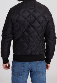 Veste bomber matelassée noire avec col, poignets et ourlet en tricot côtelé, associée à un jean en denim bleu. Le design présente une texture lisse.