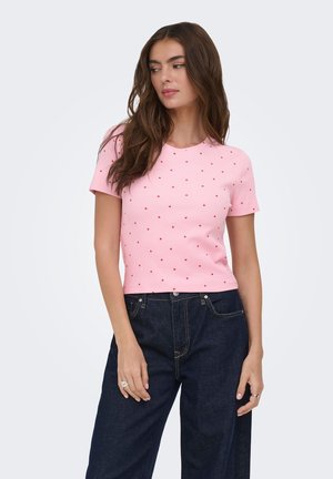 Femme aux longs cheveux bruns portant un haut rose à manches courtes avec un motif de cœurs rouges et un jean taille haute bleu foncé, debout devant un fond blanc.