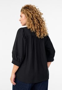 Svart, löst sittande blus med puffade ärmar, ribbad detaljering på axlarna och rundad fåll. Tillverkad av lättviktigt tyg.