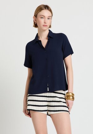 Donna con camicia blu marina a maniche corte con bottoni, pantaloncini a righe bianche e nere in maglia e braccialetti dorati spessi al polso.