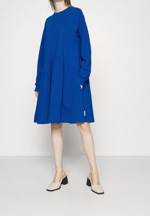 Modrá dlouhá šaty s kulatým výstřihem a nabíranou spodní částí. Pár světle béžových bot s otevřenou patou a blokovým podpatkem.