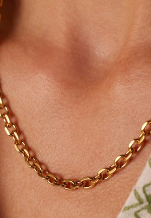 Gouden kettingbedel met ovale schakel, gepolijste afwerking, nauwsluitend om de nek, rustend op een gestructureerde huid met warme ondertonen.
