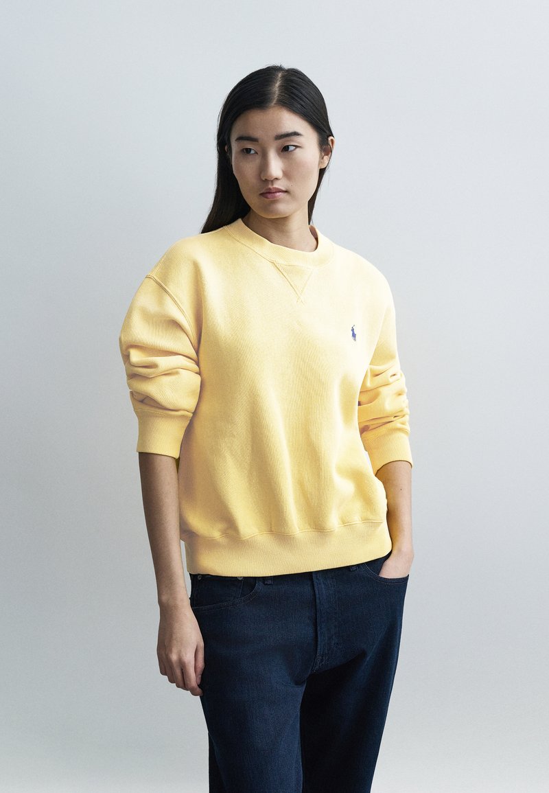 Sweat-shirt en coton jaune avec encolure ronde et poignets côtelés, arborant un petit logo bleu sur la poitrine gauche. Assorti à un jean en denim foncé.