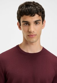 Bordeauxfarbener Strickpullover mit rundem Ausschnitt und gerippter Textur, getragen von einer Person mit kurzen, dunklen Haaren und neutralem Gesichtsausdruck.