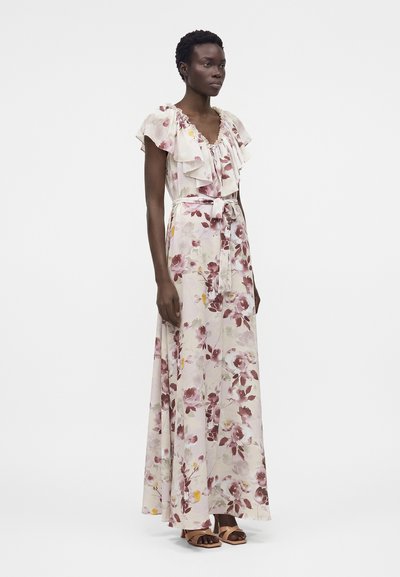 Γυναίκα που φοράει αμάνικο, μακρύ φόρεμα με floral μοτίβο, βολάν στο ντεκολτέ και ζώνη στη μέση, στέκεται μπροστά σε απλό λευκό φόντο.
