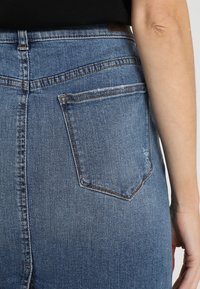 Jeans de mezclilla azul con cintura alta, detalles de costura y un único bolsillo trasero. La tela tiene una textura desgastada con un desvanecimiento sutil.