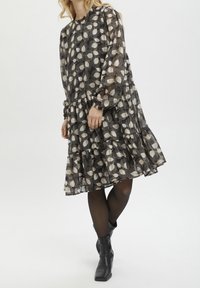 Robe noire avec motif de feuilles crème, manches longues, design à volants et poignets froncés. Portée avec des bottines noires. Tissu texturé.
