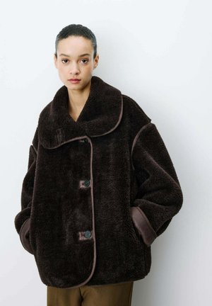 Femme portant un manteau en peluche marron foncé oversized avec grand col, boutons sur le devant et poignets bordés de cuir, debout sur fond blanc.