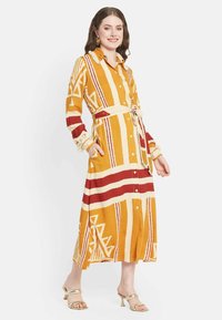 Abito button-up lungo fino alle caviglie in giallo senape con motivi geometrici rossi e bianchi. Caratterizzato da maniche lunghe, una cintura da annodare e tasche.