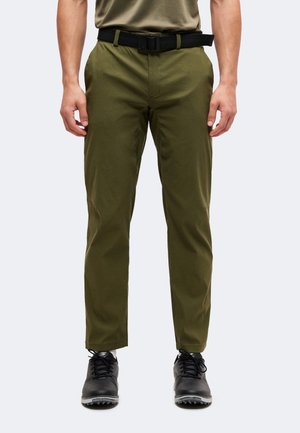 Homme portant un pantalon vert olive avec une ceinture noire et des chaussures noires, debout les mains détendues le long du corps sur un fond clair.