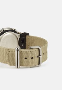 Montre avec un boîtier rond en métal et un bracelet en tissu beige. Le bracelet est doté d'une boucle en argent et de trous ajustables pour personnaliser la taille.