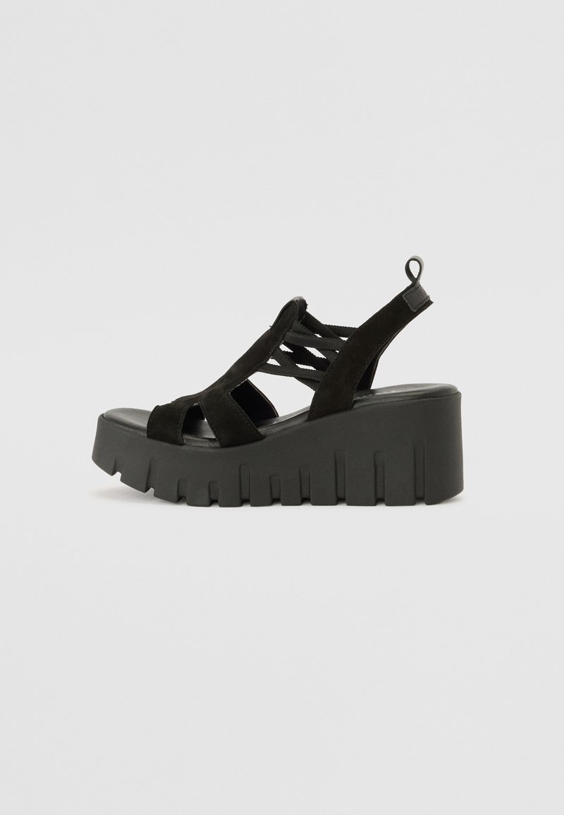 Sandale compensée noire avec semelle épaisse rainurée, bout ouvert et brides croisées avec boucle à la cheville sur fond gris uni.