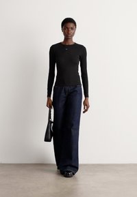 Black long-sleeve top paired with blue wide-leg jeans. The model holds a small black handbag; textured floor and plain background.