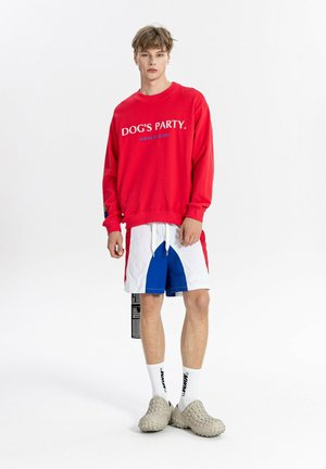 Joven con sudadera roja con el texto "DOG'S PARTY", pantalones cortos bicolor, calcetines blancos y zapatos beige sin cordones con textura.