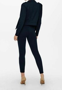 Veste courte marine avec épaulettes, associée à des leggings bleu marine moulants et des chaussures à talons hauts beige clair. Détails de texture clairs.