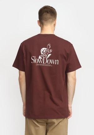 Bordeauxrotes Baumwoll-T-Shirt mit Grafik eines Schildkröte auf der Rückseite und dem Text "Langsam Machen" in Weiß, mit lässigem Schnitt und kurzen Ärmeln.