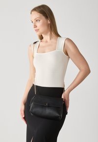 Borsa a tracolla in pelle nera con pattina ripiegata, catena dorata e dettaglio a nodo. Il modello indossa un top aderente color crema e una gonna nera.