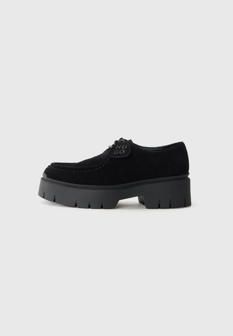 Chaussures en daim noir à lacets avec une épaisse semelle en caoutchouc noir présentant un motif nervuré. Comprend un patch rond noir avec le logo sur la languette.