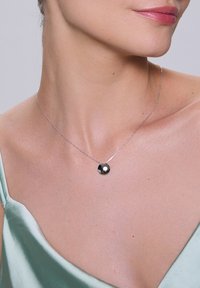 Srebrny naszyjnik z okrągłym, wypolerowanym czarnym wisiorkiem, noszony na barej skórze, z widoczną jasną zieloną satynową bluzką.