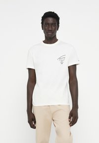 T-shirt en coton blanc avec manches courtes et col rond, présentant un logo imprimé noir sur la poitrine gauche. Texture lisse.