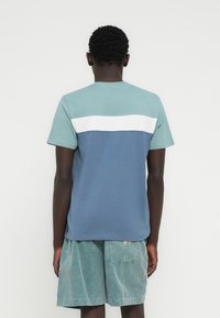 Βαμβακερό μπλουζάκι με στρογγυλή λαιμόκοψη, που διαθέτει οριζόντια μπλοκ χρωμάτων: ανοιχτό τιρκουάζ, λευκό και σκούρο μπλε. Μαλακή υφή και άνετη εφαρμογή.