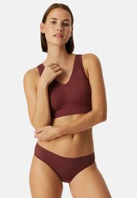 Γυναίκα με σετ μπραλέτα και εσώρουχο σε μαρόν απόχρωση, με διακριτό υφασμάτινο μοτίβο, ποζάρει με το ένα χέρι κοντά στον λαιμό και το άλλο σταυρωμένο.