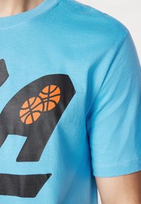 Světle modré bavlněné tričko s velkým černým grafickým designem a dvěma oranžovými ilustracemi basketbalů, s krátkými rukávy a kulatým výstřihem.