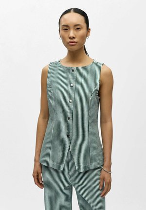 Frau mit dunklem Haar, das zu einem tiefen Pferdeschwanz gebunden ist, trägt ein ärmelloses, grün-weiß gestreiftes, geknöpftes Oberteil und passende Hose, schaut nach vorne.