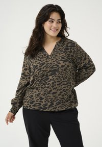 Braune Leopardenmuster-Pullover mit V-Ausschnitt und langen Ärmeln, aus einem weichen Material gefertigt. Kombiniert mit schwarzen Hosen.